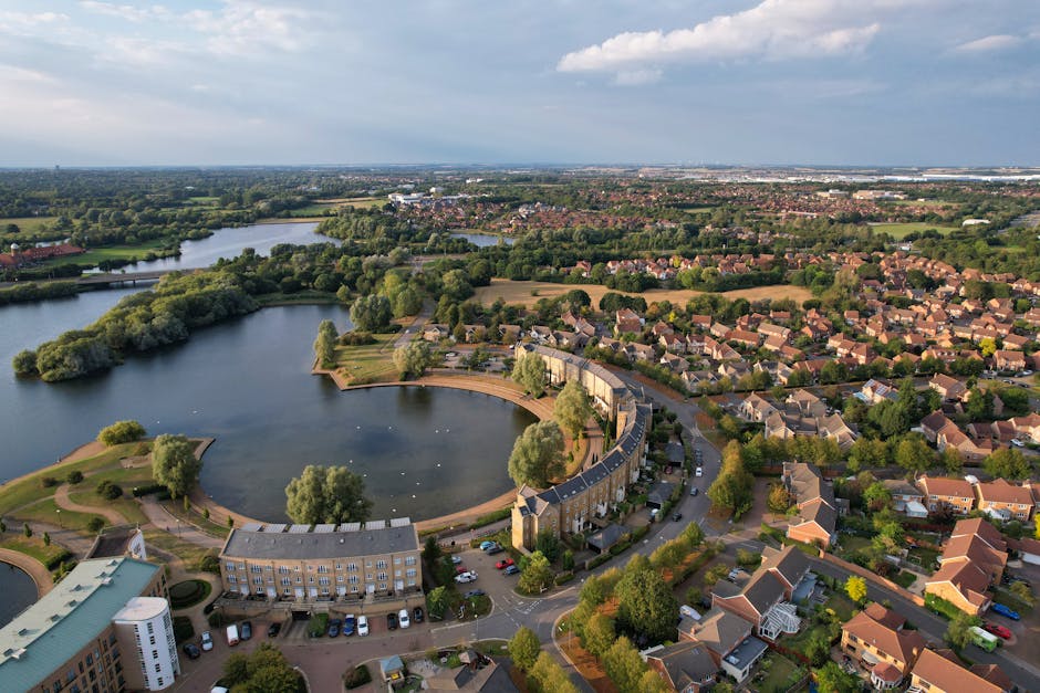 Aerial view of Arnos Park in Southgate showing a large, curved ornamental pond surrounded by a mix of residential housing and green spaces. The pond is partly encircled by a paved walkway with several trees lining the perimeter, and a small island with additional trees is visible within the water. Alongside the pond, residential buildings with flat and pitched roofs are positioned close together, with some featuring parking areas and driveways. The surrounding neighborhood consists of neatly arranged houses with gardens, trees, and streets extending into the distance, under a partly cloudy sky with patches of sunlight highlighting the landscape. The image captures the peaceful, green environment typical of home relocation and moving processes in the Southgate area, with a focus on community and natural scenery, reflecting the context of house removals and furniture transport. Man With a Van Southgate provides local removals services, including packing and loading within such residential settings.
