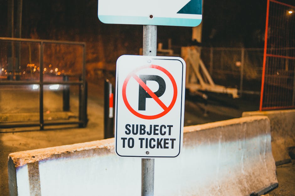 A close-up image of a white rectangular parking prohibition sign with a red circle and slash over a black letter 'P,' indicating no parking. The sign also displays the text 'SUBJECT TO TICKET' in bold black letters. It is mounted on a metal pole situated behind a concrete parking barrier at night, with warm street lighting illuminating the scene. In the background, there is a chain-link fence, some equipment or scaffolding, and a blurred view of additional road or construction area. The image is relevant to house removals and parking restrictions, which may impact the logistics of furniture transport and home relocation services offered by Man With a Van Southgate.
