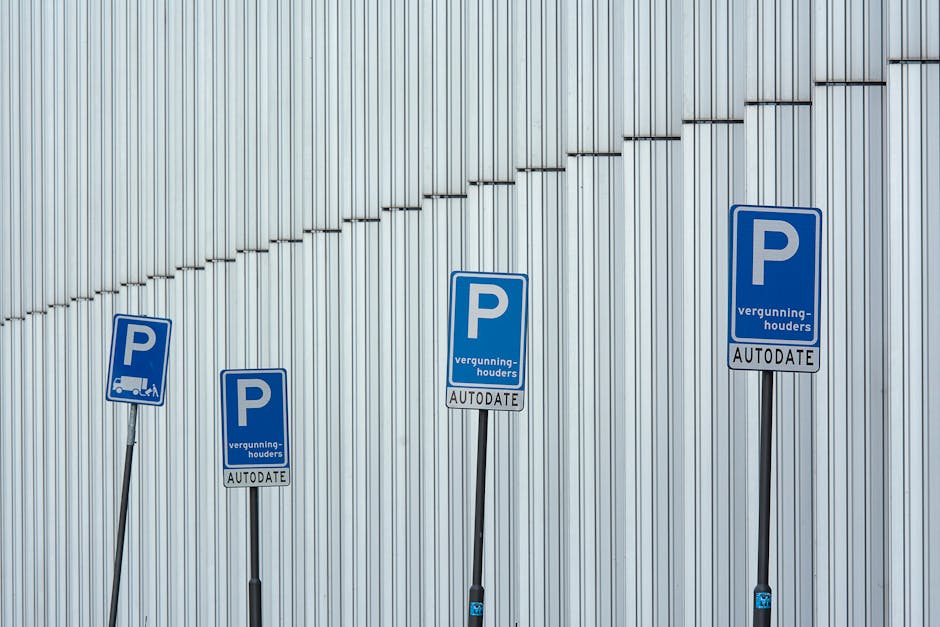 Four blue parking permit signs mounted on black poles are positioned in front of a large, vertically paneled metallic wall. The signs display a white 'P' symbol and contain text indicating parking permissions with the words 'vergunning-houders' and 'AUTODATE' printed on them. The signs are arranged in a row from left to right, at varying heights, with the lowest on the left and the highest on the right. The metallic wall behind features a pattern of evenly spaced vertical ridges and a diagonal line created by a series of small protrusions or decorative elements. The scene is lit evenly, emphasizing the metallic texture and the clarity of the signage, suggesting an outdoor or semi-enclosed urban environment related to parking regulation for vehicle access or loading zones, which is relevant to house removals and moving services discussed on the page [PAGE_TITLE] at manwithavansouthgate.co.uk.