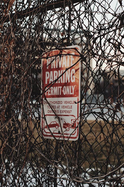 A close-up image of a white rectangular parking prohibition sign with a red circle and slash over a black letter 'P,' indicating no parking. The sign also displays the text 'SUBJECT TO TICKET' in bold black letters. It is mounted on a metal pole situated behind a concrete parking barrier at night, with warm street lighting illuminating the scene. In the background, there is a chain-link fence, some equipment or scaffolding, and a blurred view of additional road or construction area. The image is relevant to house removals and parking restrictions, which may impact the logistics of furniture transport and home relocation services offered by Man With a Van Southgate.