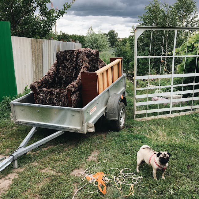 A small metal utility trailer parked on a grassy outdoor area near a white wooden fence and a garden trellis. The trailer is loaded with a large, irregularly shaped piece of bark or wood and a partially visible wooden furniture panel, secured with straps. In the foreground, a small pug dog with a pink collar stands on the grass, surrounded by loose orange and white ropes. The scene is set outdoors on a cloudy day, with trees and shrubs in the background, and an overall context of home relocation, furniture transport, or packing and moving process supported by Man With a Van Southgate, viewed from the side and capturing the loading or unloading of bulky items as part of a house removal service.
