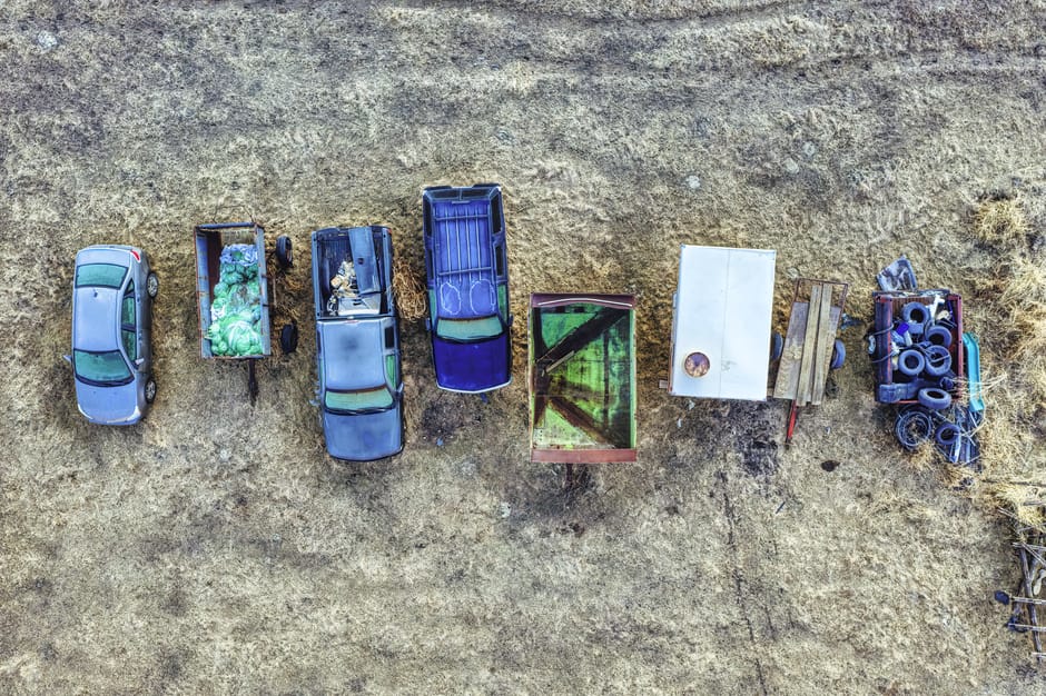 An aerial view showing seven miniature vehicles arranged in a line on a patch of dry, brown grass, positioned from left to right: a grey car, a small open-top trailer filled with green waste and recyclables, a dark blue van with a partially open rear door revealing some cargo, a bright blue truck with an open cargo area, a green and red painted wooden box, a white rectangular table top, and a small blue utility trailer loaded with black rubber tires and coiled hoses. The scene takes place outdoors in daylight, with the vehicles aligned as if part of a furniture transport or home relocation process. The vehicles appear to be scale models, but are detailed with realistic textures and colors that suggest transportation and packing activities related to house removals. Man With a Van Southgate specializes in removals and moving services, which is subtly reflected in the arrangement of these objects and their context within the image.