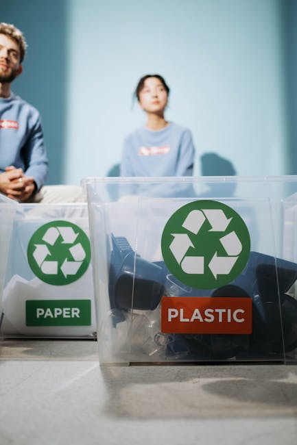 Inside a room with a light blue wall, two individuals are seated behind a table that holds clear plastic recycling bins. The bin on the left is labeled 'PAPER' and contains crumpled paper, while the bin on the right is labeled 'PLASTIC' and holds various plastic items such as bottles and caps. The bins feature green recycling symbols. In the background, a man with dark hair and a beard wearing a blue top and a woman with dark hair in a ponytail, both dressed in light blue tops, are visible but slightly out of focus. The scene is well-lit and captures a moment related to eco-friendly waste separation, which aligns with environmentally conscious aspects of house removals and packing practices conducted by Man With a Van Southgate during home relocation and furniture transport processes.