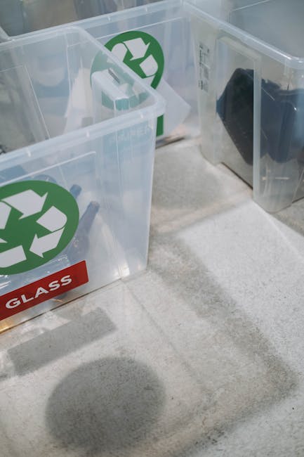 Inside a home visible in this image are three clear plastic recycling and waste containers, each marked with green recycling symbols and specific labels for glass disposal. The containers are positioned on a light-colored carpeted floor near a doorway, with one container partly obscured. The containers are empty, with some small pieces of packaging or paper inside. The scene appears to be part of a home moving or packing process coordinated by Man With a Van Southgate, which may involve sorting waste and recycling materials during house removals or furniture transport preparations, illustrating the logistical steps in packing and moving services. The natural lighting highlights the transparency of the containers and the labels, emphasizing a focus on recycling and eco-friendly disposal practices during a home relocation.