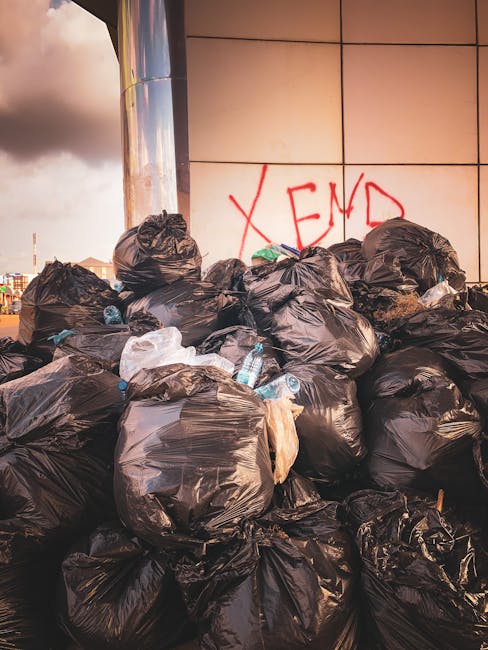 A pile of tightly packed black rubbish bags filled with household waste, including discarded plastic bottles and packaging, situated outdoors against a building wall with beige tiles. The bags are sta