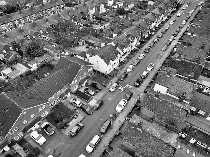 An aerial black-and-white photograph showing a residential street in Southgate with closely packed terraced houses and semi-detached homes. The street is lined with parked cars on both sides, including sedans and small SUVs, with some vehicles occupying spaces on the curb. A van from Man With a Van Southgate is parked on the pavement, partially blocking the entrance to a house, which is being prepared for a home relocation or furniture transport. The pavement is clear except for the van and a small trolley nearby. Tucked between the houses are narrow driveways, some with additional parked cars, and small backyard gardens. The rooftops of the houses feature tiles or shingles, with varying heights and orientations, and a few chimneys are visible. Street lighting fixtures and traffic markings are seen along the road, emphasizing the urban environment suitable for local moving logistics and packing and moving activities, consistent with the services offered by Man With a Van Southgate.