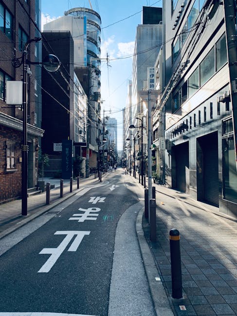 A narrow urban street scene with tall modern glass and concrete office buildings on both sides, featuring a one-way road with white directional arrows and Japanese characters painted on the asphalt. The street is mostly empty, lined with black bollards and street lamps, with overhead power lines and cables visible overhead. The weather is clear with a blue sky and some clouds. This setting illustrates an area suitable for home relocations or furniture transport in a dense city environment, relevant to house removal services like those provided by Man With a Van Southgate near Chase Side, Southgate, in challenging narrow street conditions.