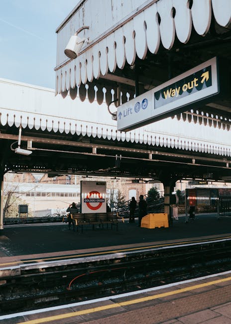 The image depicts the exterior of Golders Green underground station with train tracks in the foreground and a covered platform area overhead. The platform features wooden benches for waiting passenger