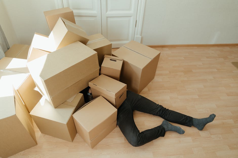 A person lying face down on a wooden floor inside a room, partially hidden beneath a large pile of unpacked cardboard boxes, some with handles and others plain, varying in size and stacked haphazardly. The boxes occupy the center and left side of the image, with natural light illuminating the scene. The person is wearing black pants and grey socks, with their legs extended outward. Behind the scene, there's a white door and a beige wall, indicating a typical interior space. The arrangement suggests ongoing packing or moving activities as part of a home relocation process, consistent with furniture transport and packing and moving services offered by Man With a Van Southgate. The clutter of boxes and the visible effort involved in packing reflect the logistical challenges of last-minute removals, aligning with themes of packing panic and move survival. The image captures the typical environment encountered during house removals, emphasizing the physical demands and clutter involved in a late-stage move inside a residential property.