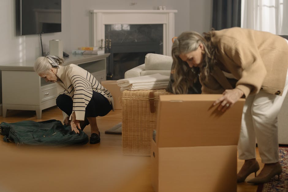 A woman with grey hair, wearing a striped long-sleeve shirt and black pants, is kneeling on the wooden floor, packing a green backpack with a focused expression. Next to her, another woman with long grey hair, dressed in a beige coat and white pants, is bent over, lifting or adjusting a large cardboard box. The scene takes place inside a well-lit living room equipped with a white fireplace, a white entertainment unit with a mounted TV, and a grey sofa with folded towels or sheets placed on its armrest. The floor is wooden, and natural light filters through sheer curtains. Nearby, a wicker basket and other moving packing materials are visible. This image captures the process of home relocation, including packing, furniture transport, and loading activities, which are part of the services provided by Man With a Van Southgate, supporting last-minute move preparation and interior packing.