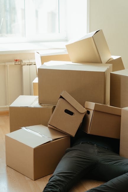 A person lying face down on a wooden floor inside a room, partially hidden beneath a large pile of unpacked cardboard boxes, some with handles and others plain, varying in size and stacked haphazardly. The boxes occupy the center and left side of the image, with natural light illuminating the scene. The person is wearing black pants and grey socks, with their legs extended outward. Behind the scene, there's a white door and a beige wall, indicating a typical interior space. The arrangement suggests ongoing packing or moving activities as part of a home relocation process, consistent with furniture transport and packing and moving services offered by Man With a Van Southgate. The clutter of boxes and the visible effort involved in packing reflect the logistical challenges of last-minute removals, aligning with themes of packing panic and move survival. The image captures the typical environment encountered during house removals, emphasizing the physical demands and clutter involved in a late-stage move inside a residential property.