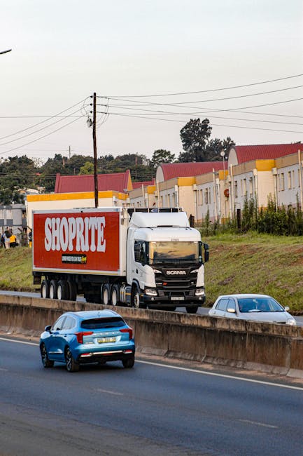 A white Scania box truck with a red 'SHOPRITE' logo on its side is driving on a multi-lane road, with a smaller blue car and a white car visible in the foreground. The truck is positioned on the upper level of the road, which appears to be a highway or busy street, with a slight incline. In the background, there are residential buildings with red roofs and a row of trees, under an overcast sky. Utility poles and power lines run parallel to the road. This scene depicts a typical home relocation or furniture transport process involving professional removals, such as those by Man With a Van Southgate, with vehicles used for moving household items efficiently across urban routes.