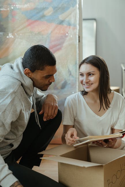 Inside a residential property, a male mover from Man With a Van Southgate is assisting with home relocation by handling a cardboard box filled with household items, while a woman, holding a notebook o