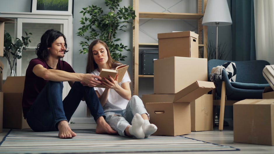 Inside a residential property, a male mover from Man With a Van Southgate is assisting with home relocation by handling a cardboard box filled with household items, while a woman, holding a notebook o