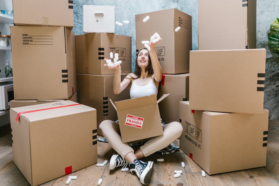 A young woman with long dark hair, seated on a wooden floor inside a room, surrounded by large cardboard moving boxes of various sizes, some sealed with red packing tape and others open. She is wearing a white tank top, beige pants, and black and white sneakers, and is smiling while tossing packing foam into the air. The open box around her contains foam packing peanuts, with additional packing materials scattered on the floor. Behind her, the walls are painted in a light blue and textured finish, and a green potted plant is partially visible on the right side of the image. The scene captures a home relocation or furniture transport context, with the woman expressing a moment during packing or unpacking as part of a house move, reflecting the logistics involved in house removals supported by Man With a Van Southgate.