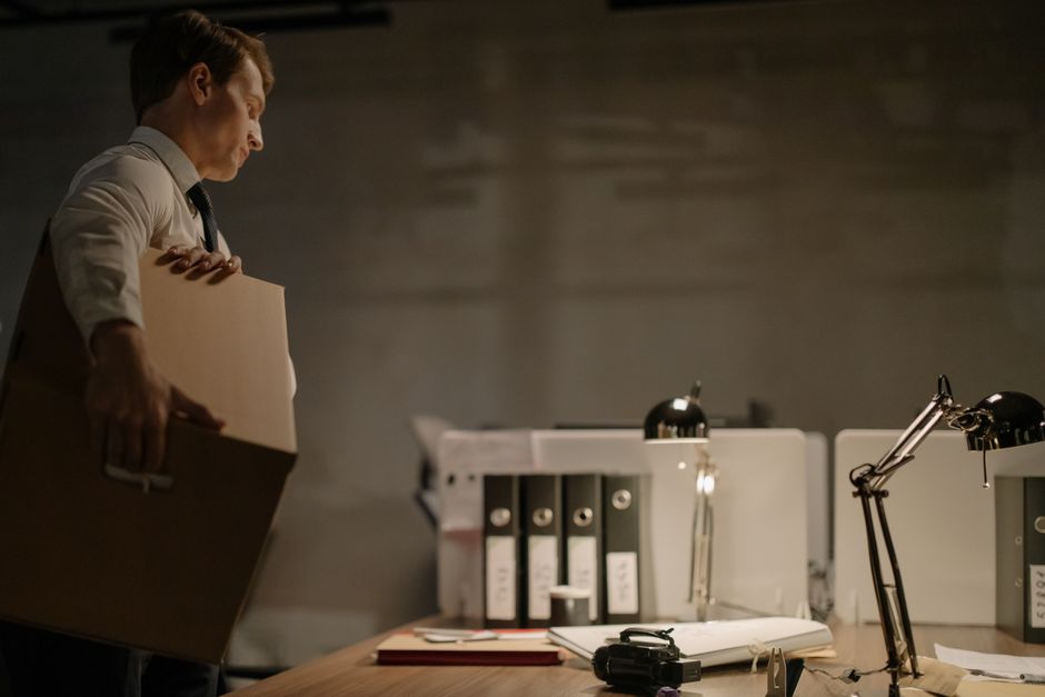 A professional man with light skin and short brown hair, wearing a light-colored shirt and dark tie, is seen carrying a large cardboard box in an office environment. The office features a wooden desk on which there are black and white ring binders, a set of paperwork, a camera, and two adjustable black desk lamps. The background includes a gray wall with a textured finish. The scene captures a moment during a packing and moving process, illustrating the logistics involved in office furniture transport and home relocation services provided by Man With a Van Southgate, which may include packing, loading, and transporting items such as office furniture, documents, and equipment.