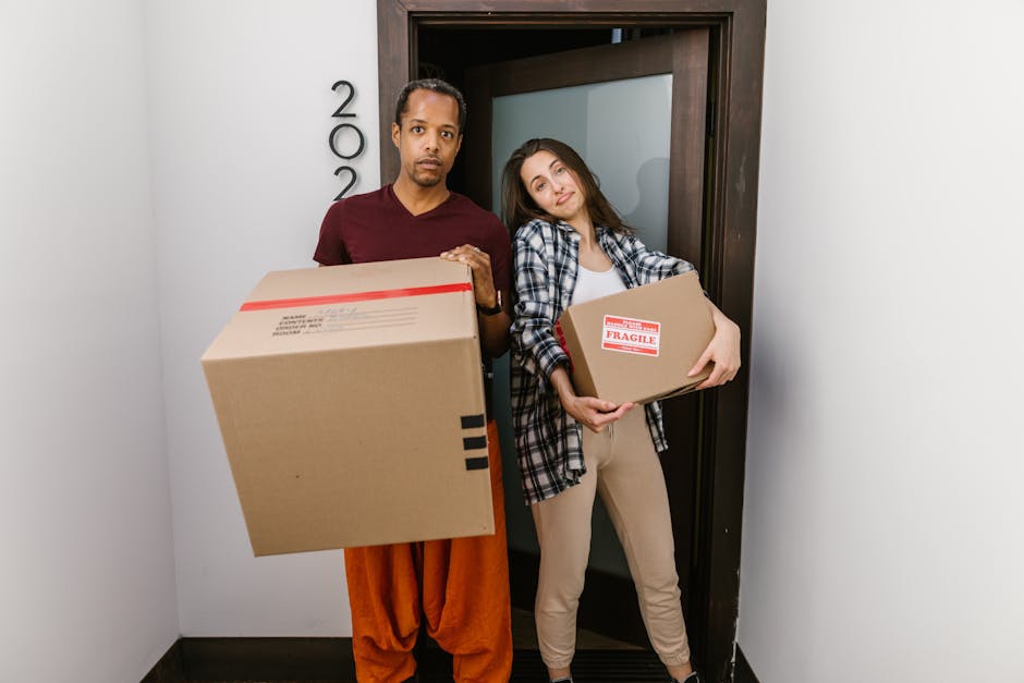 A young woman with long dark hair, seated on a wooden floor inside a room, surrounded by large cardboard moving boxes of various sizes, some sealed with red packing tape and others open. She is wearing a white tank top, beige pants, and black and white sneakers, and is smiling while tossing packing foam into the air. The open box around her contains foam packing peanuts, with additional packing materials scattered on the floor. Behind her, the walls are painted in a light blue and textured finish, and a green potted plant is partially visible on the right side of the image. The scene captures a home relocation or furniture transport context, with the woman expressing a moment during packing or unpacking as part of a house move, reflecting the logistics involved in house removals supported by Man With a Van Southgate.