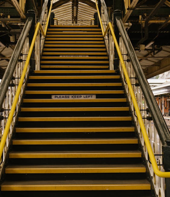 Indoor view of a metal staircase with yellow handrails leading up to an upper level within a building, possibly a warehouse or industrial site. The stairs are made of dark material with yellow edge markings on each step and a white sign with black lettering that reads 'PLEASE KEEP LEFT' affixed to one of the lower steps. The environment includes visible structural elements like beams and pipes overhead, illuminated by industrial lighting. This staircase may be part of a location used for loading or moving heavy furniture or equipment during home relocation or furniture transport, as seen in professional relocations by companies such as Man With a Van Southgate.
