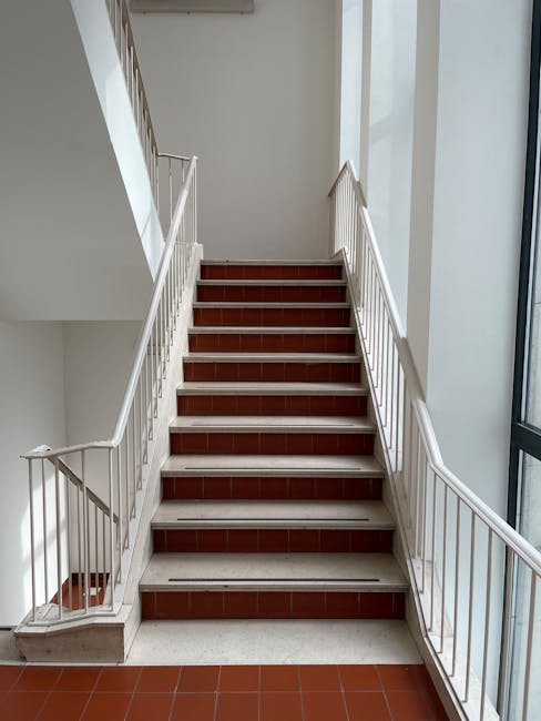A straight staircase inside a residential building with white metal handrails on each side, leading upwards to an upper floor. The stairs are covered with reddish-brown tiles on each step, and there is a narrow gap between the floor and the first step. To the right of the staircase, there is a large floor-to-ceiling window allowing natural light to illuminate the area. The staircase is situated adjacent to a plain white wall, and the surrounding environment suggests an indoor communal space, possibly in a flat or apartment block. The setting is clean and well-maintained, typical of a professional home relocation environment where furniture and packing materials may be moved through this area as part of the services provided by Man With a Van Southgate, specialists in removals and moving logistics.