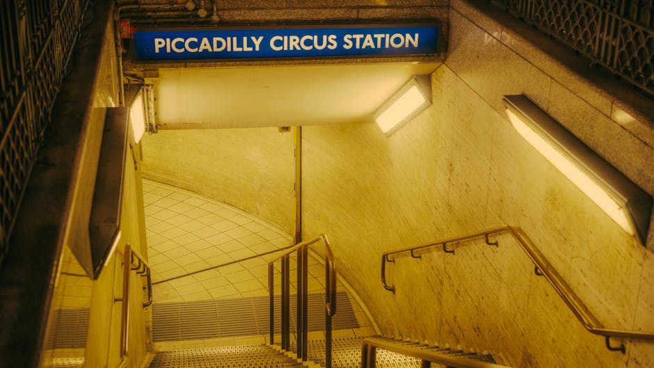 Indoor view of a metal staircase with yellow handrails leading up to an upper level within a building, possibly a warehouse or industrial site. The stairs are made of dark material with yellow edge markings on each step and a white sign with black lettering that reads 'PLEASE KEEP LEFT' affixed to one of the lower steps. The environment includes visible structural elements like beams and pipes overhead, illuminated by industrial lighting. This staircase may be part of a location used for loading or moving heavy furniture or equipment during home relocation or furniture transport, as seen in professional relocations by companies such as Man With a Van Southgate.