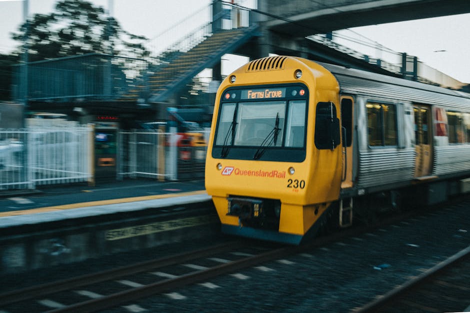 A yellow Queensland Rail train traveling along train tracks at an urban station, with a digital destination sign displaying 'Ferry Grove' on the front. The train is a modern electric model, with a streamlined design and multiple windows along its side. In the background, there is a blue pedestrian footbridge with stairs, metal railings, and some pedestrians crossing. The platform area includes a yellow safety line along the edge and there are various equipment and signage visible nearby. The scene is lit by natural daylight, indicating daytime, and captures a typical urban railway environment that could be part of a house relocation or transport logistics context, relevant to house removals and moving services offered by Man With a Van Southgate.