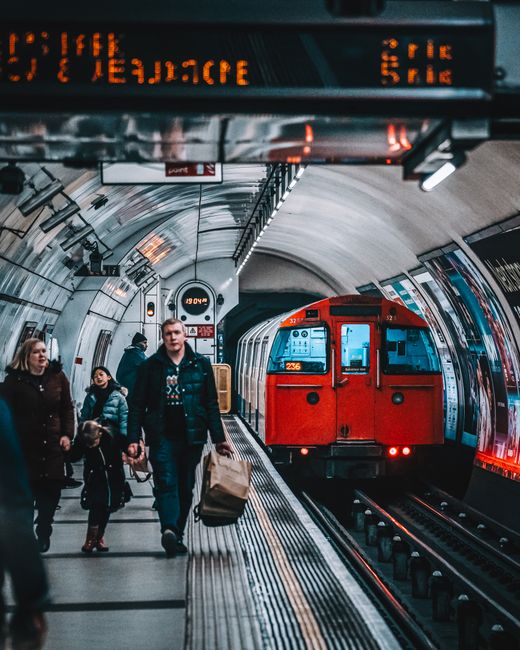 A group of people, including a man carrying a paper bag, walk along an underground train station platform with a red train approaching in the background. The station features a curved ceiling with metallic panels, illuminated signs, and electronic displays overhead showing train times and destination information. The platform has tactile paving near the edge, and several other passengers are seen waiting or walking nearby. This scene depicts the transportation aspect of house removals and home relocation services, where a professional team from Man With a Van Southgate may coordinate travel logistics for furniture transport or a move between residences.